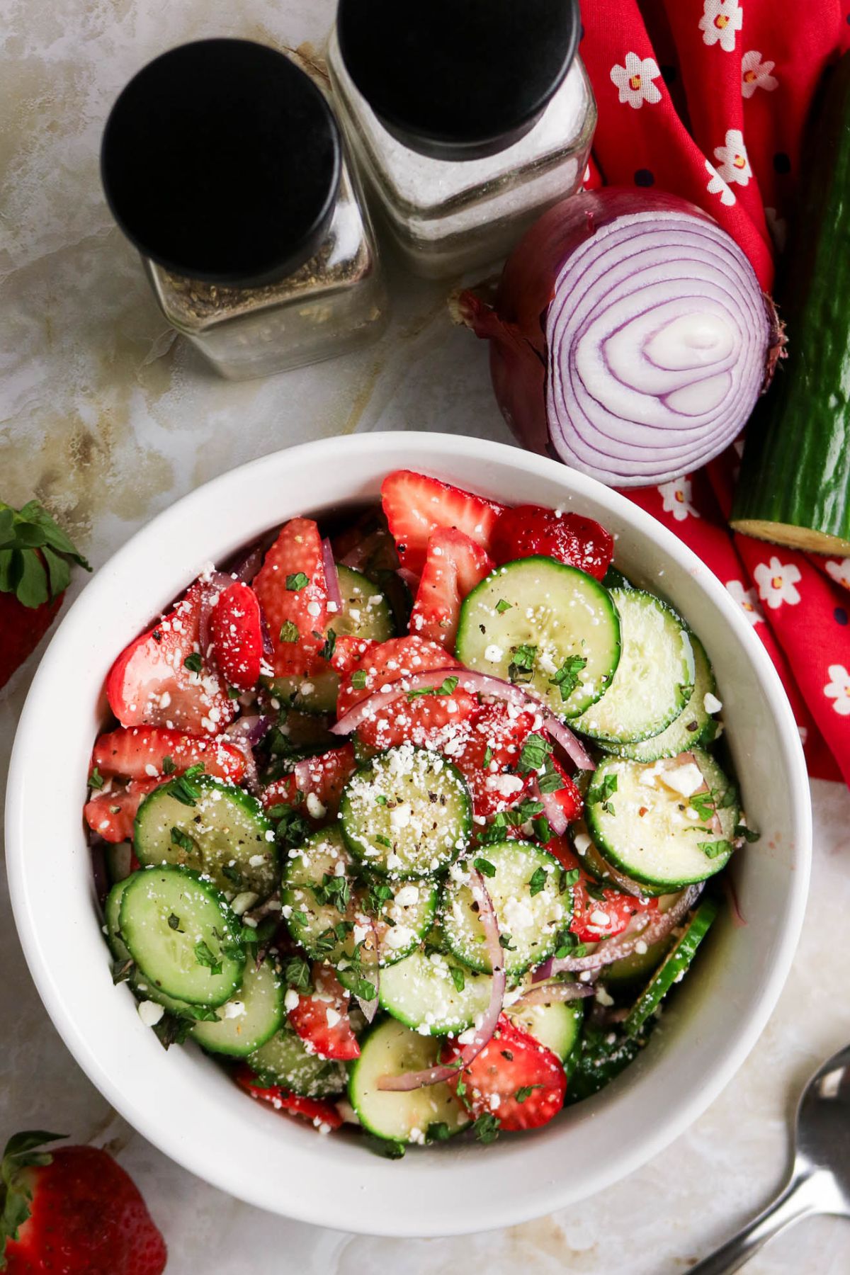 A bowl of salad with sliced cucumbers, strawberries, red onion, herbs, and crumbled cheese. Salt and pepper shakers, a red onion, and a red cloth are nearby.
