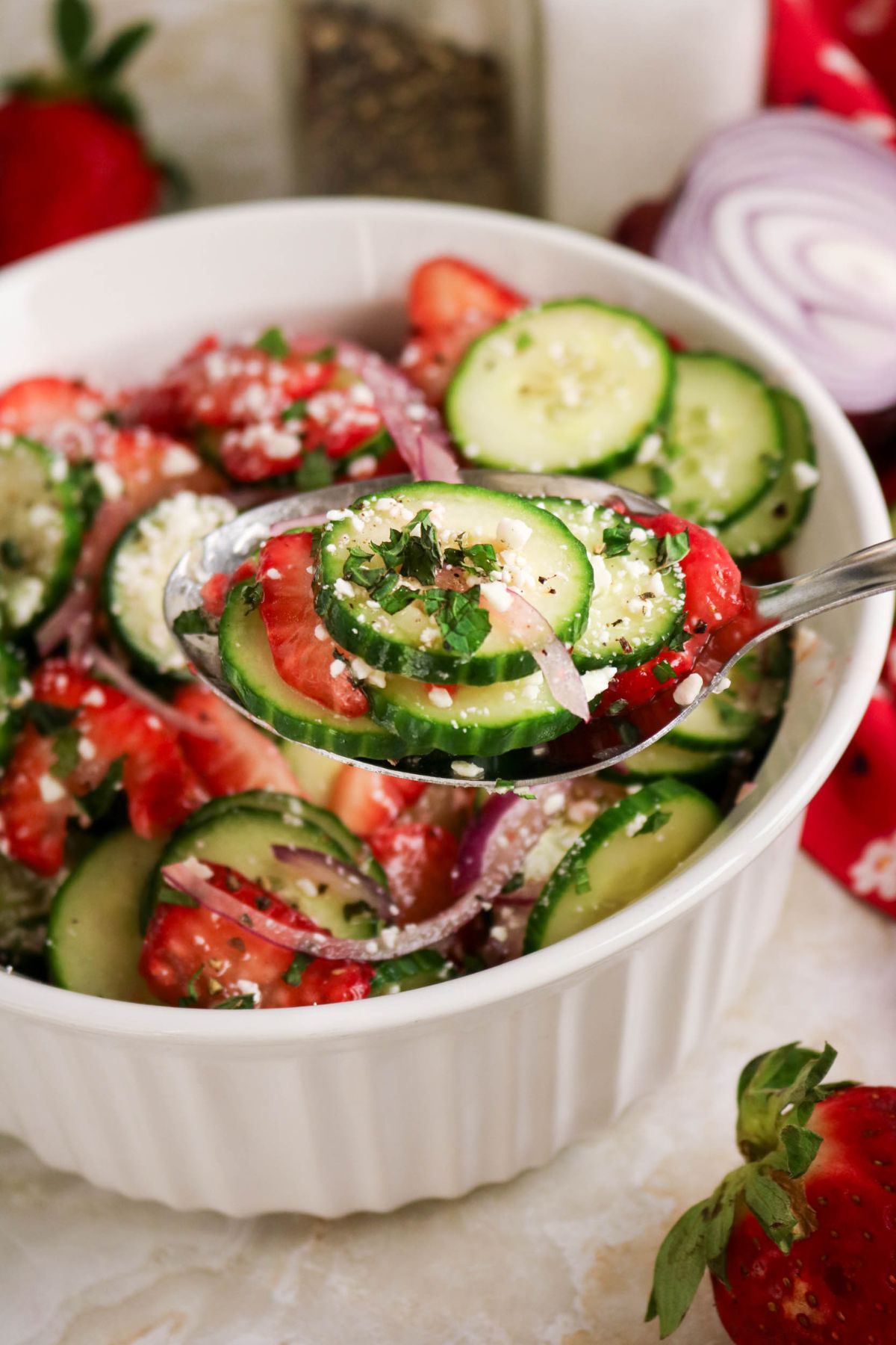 A spoonful of cucumber, strawberry, red onion, and feta salad being lifted from a white bowl, with herbs sprinkled on top.