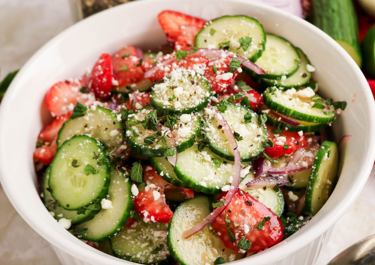 A bowl of salad with sliced cucumbers, strawberries, red onions, crumbled cheese, and herbs.