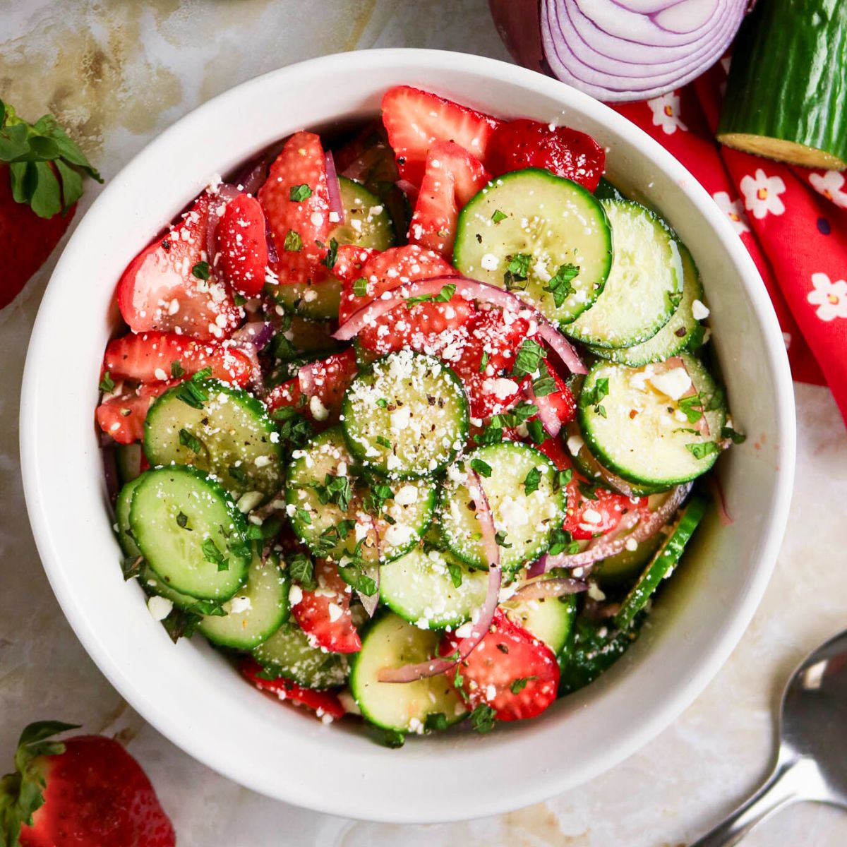 A bowl of salad containing sliced cucumbers, strawberries, red onions, herbs, and a sprinkle of cheese on top.