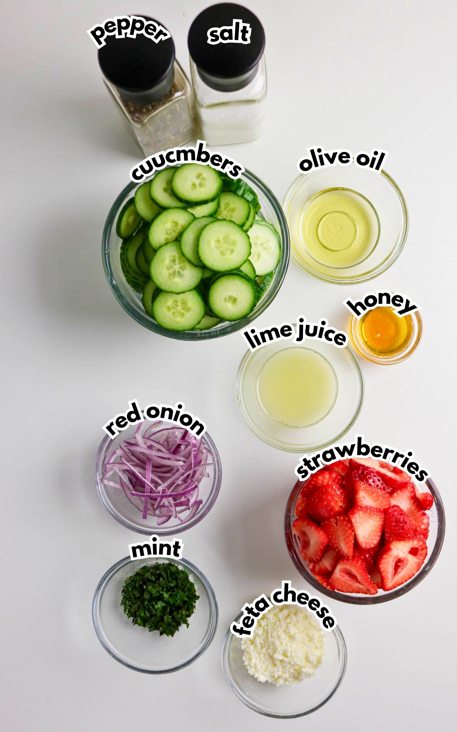Ingredients for a salad displayed in bowls on a white surface: cucumbers, strawberries, red onion, mint, feta cheese, olive oil, lime juice, honey, salt, and pepper.