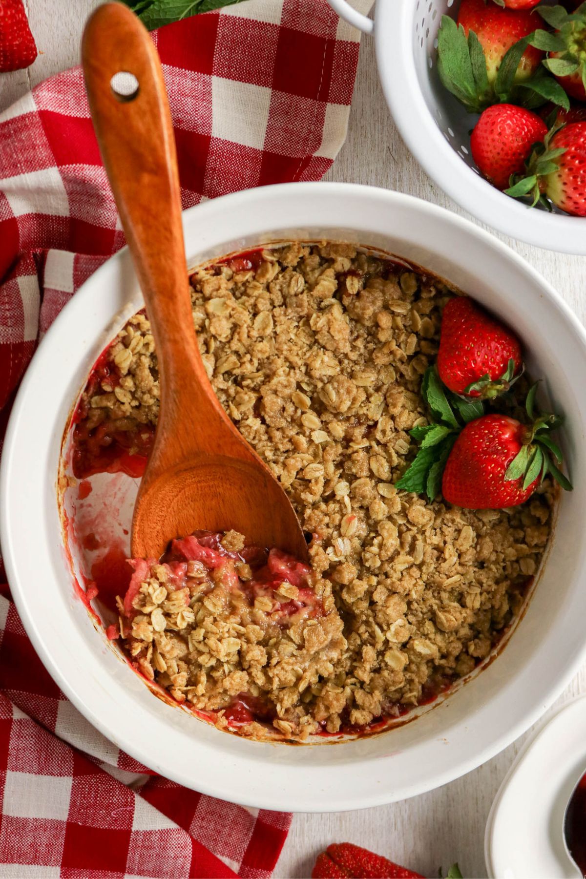 A baking dish of strawberry crisp with oats topping, partially served with a wooden spoon, sits on a red and white checkered cloth next to fresh strawberries.