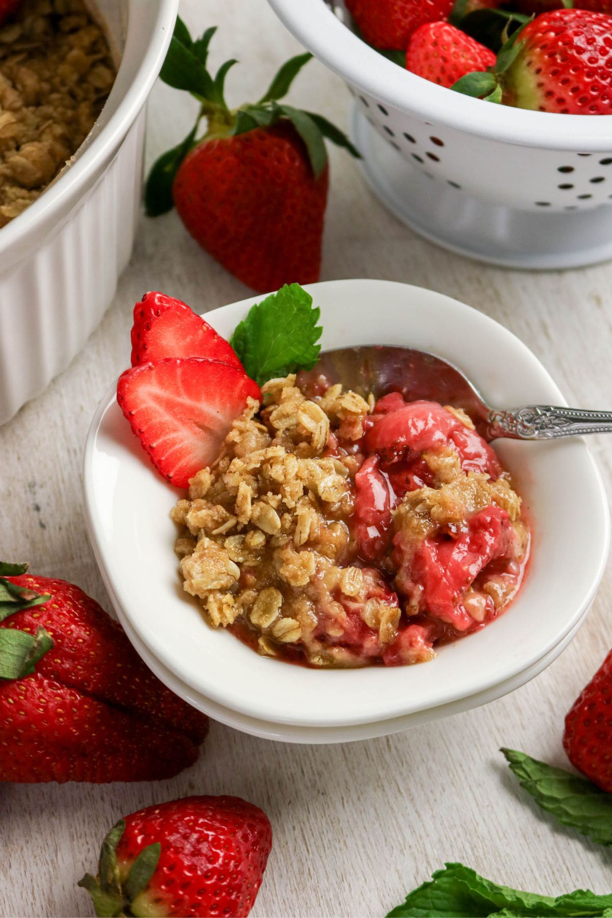 A bowl of strawberry crisp topped with oat crumble, garnished with fresh strawberry slices and a mint leaf, surrounded by whole strawberries.