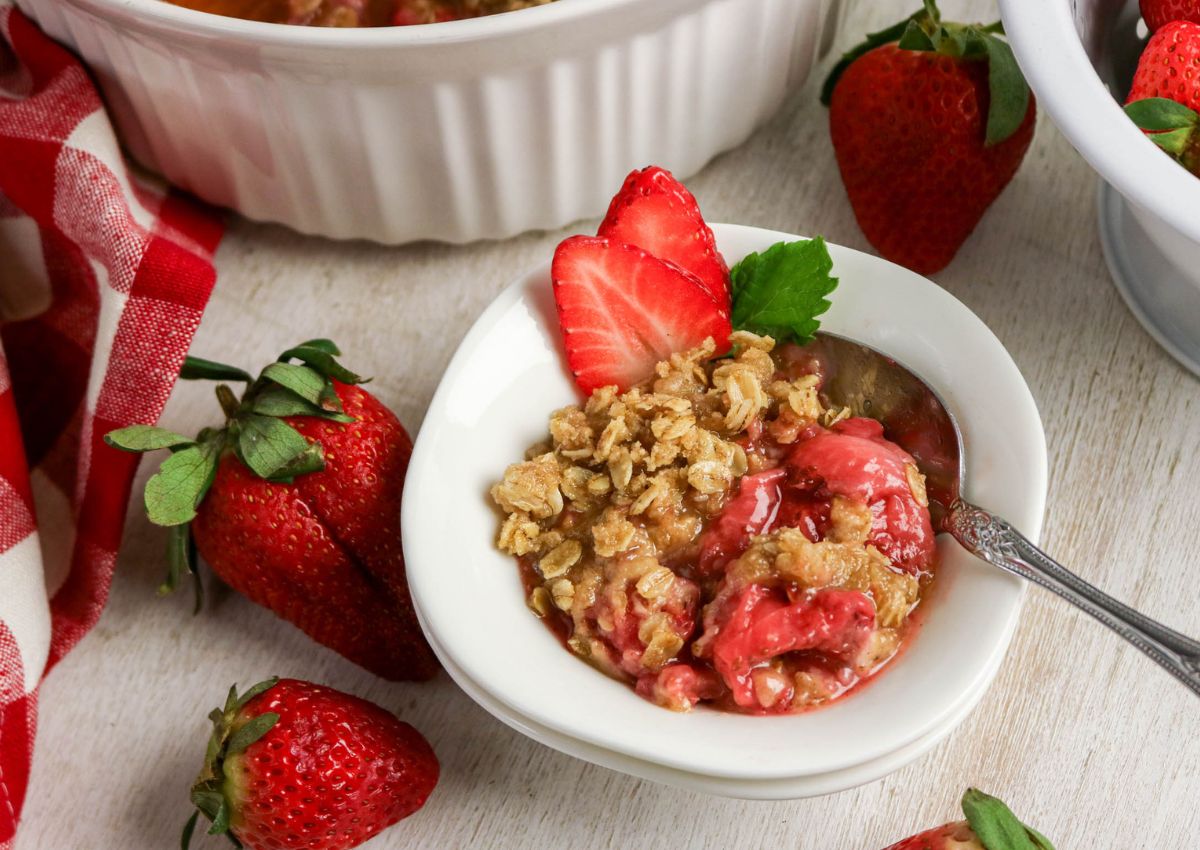 A bowl of strawberry crumble topped with oat streusel and a fresh strawberry slice, with whole strawberries and a red checkered cloth nearby.