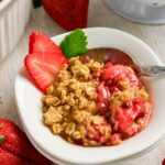A bowl of strawberry crisp topped with oat crumble, garnished with two strawberry slices and a green mint leaf, with a spoon in the bowl.