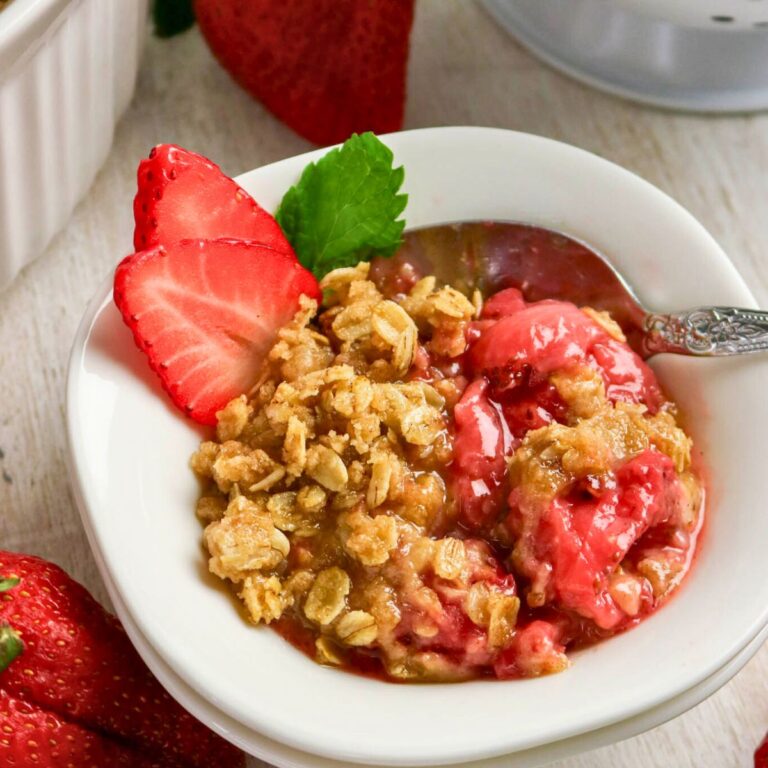 A bowl of strawberry crisp topped with oat crumble, garnished with two strawberry slices and a green mint leaf, with a spoon in the bowl.