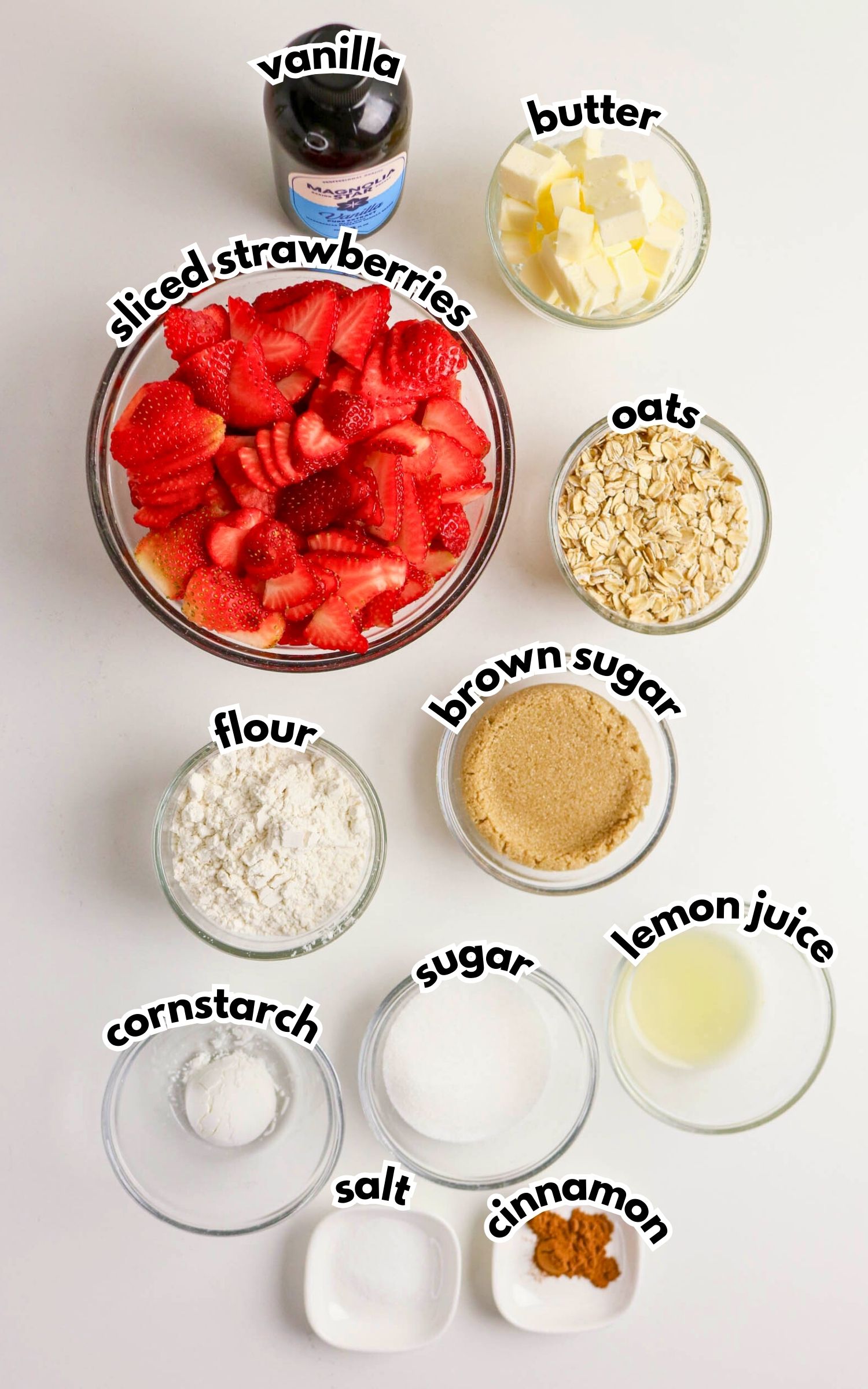 Overhead view of labeled ingredients for baking, including sliced strawberries, vanilla, butter, oats, brown sugar, flour, cornstarch, sugar, salt, lemon juice, and cinnamon.