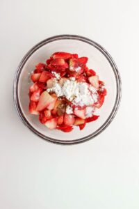 A glass bowl filled with chopped strawberries and rhubarb topped with a layer of white sugar and cornstarch on a white surface.