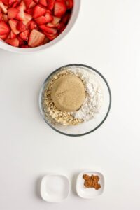 A bowl of sliced strawberries, a bowl with brown sugar, flour, and oats, and two small dishes containing sugar and ground spices on a white surface.
