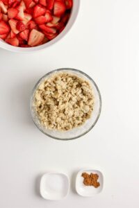 A bowl of sliced strawberries, a bowl of oat crumble topping, and two small dishes containing salt and ground cinnamon on a white surface.