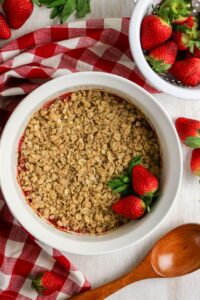 A round baking dish of strawberry crisp with oat topping, garnished with fresh strawberries, on a red-and-white checkered cloth next to a wooden spoon and a bowl of strawberries.
