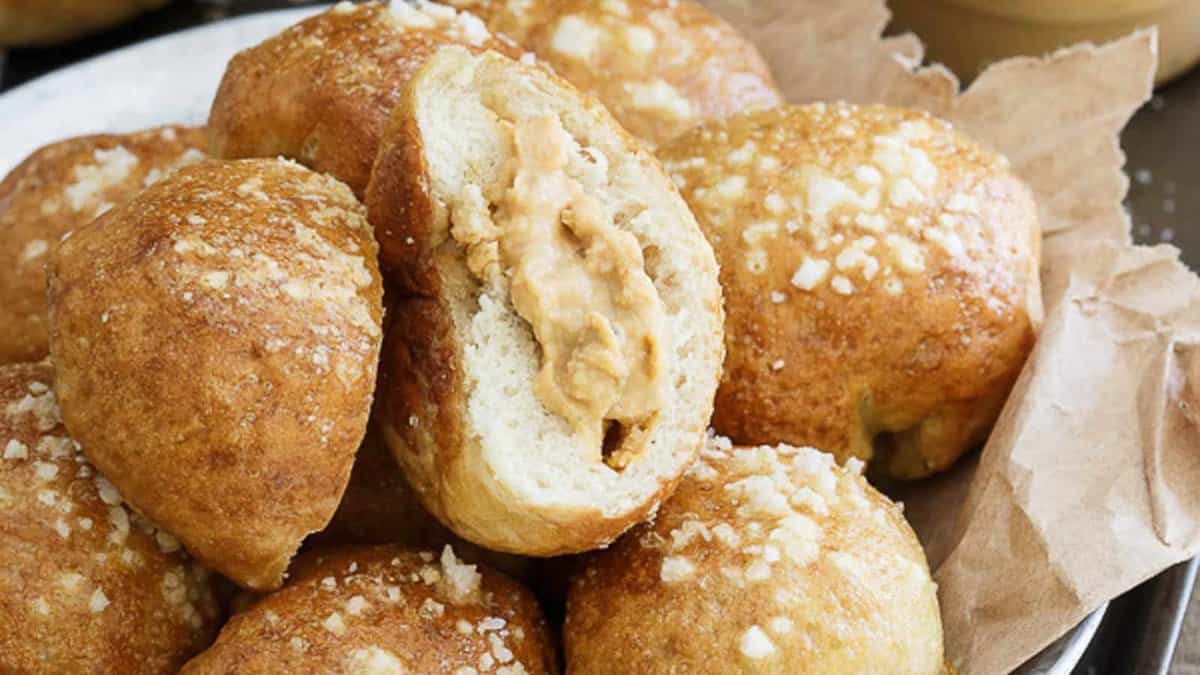 A close-up of several round bread rolls topped with coarse sugar, with one roll split open to show a creamy peanut butter filling inside.