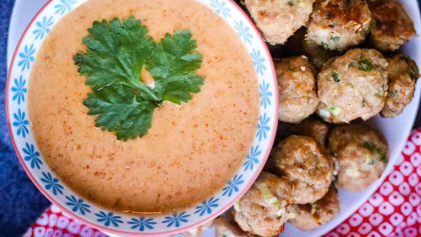 A bowl of creamy orange dipping sauce garnished with cilantro, next to a plate of small, round meatballs.
