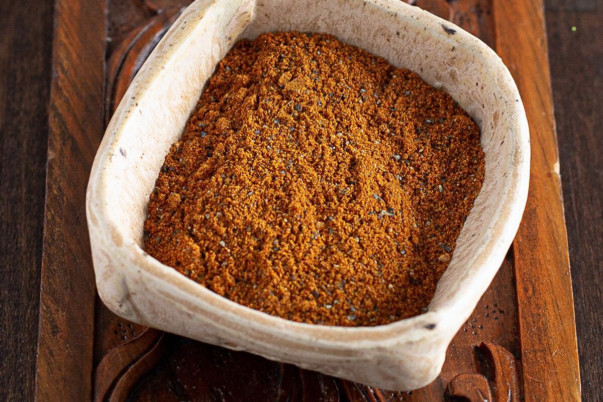 A square ceramic bowl filled with a reddish-brown ground spice blend sits on a carved wooden surface.