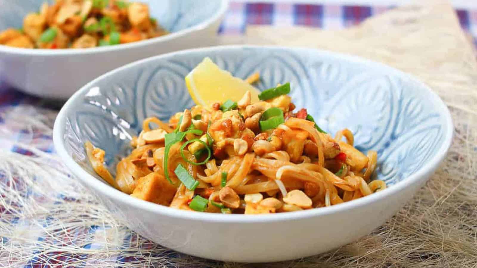 A bowl of Pad Thai noodles garnished with chopped peanuts, green onions, and a lemon wedge, placed on a patterned surface.