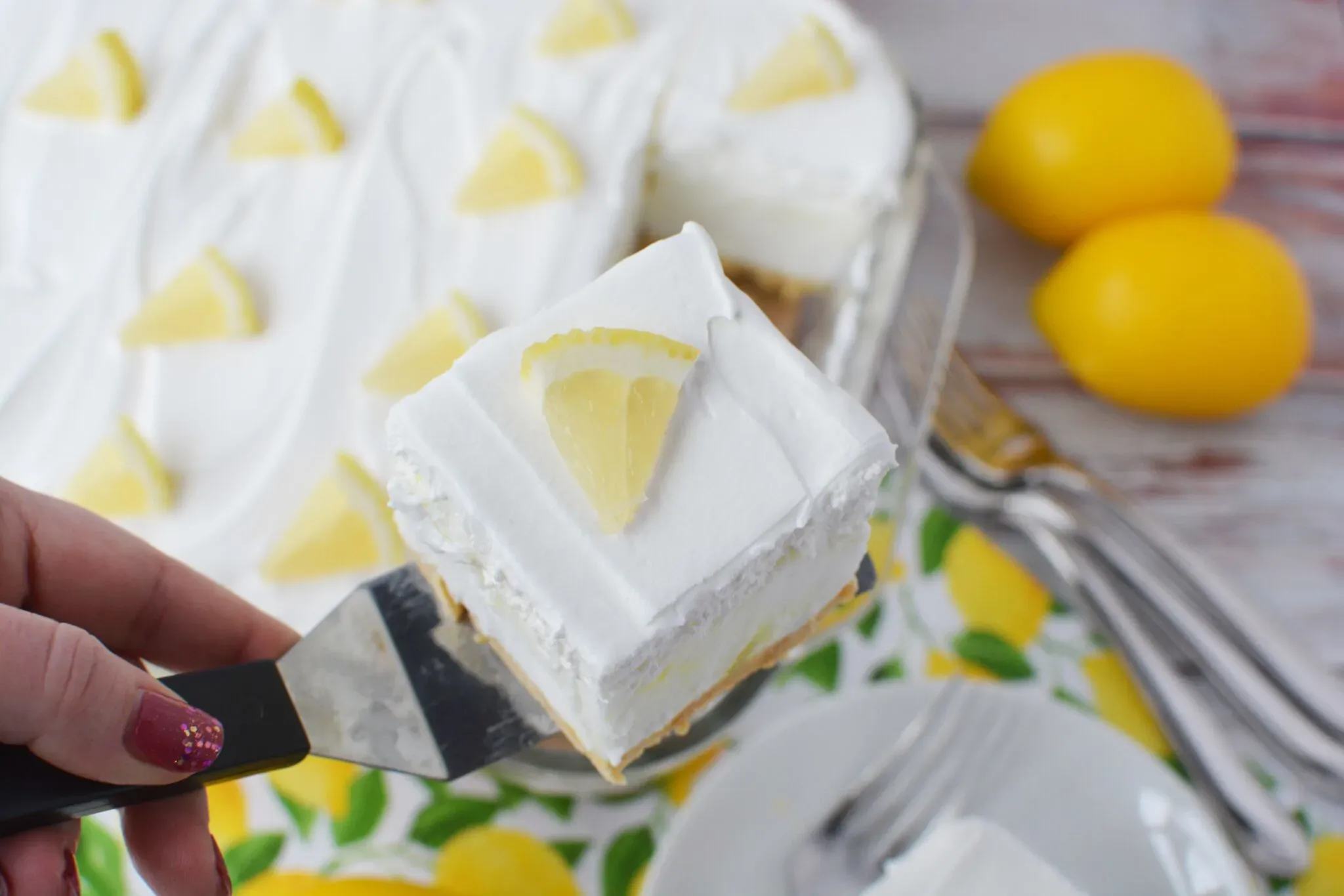 A hand holds a spatula with a square slice of lemon dessert topped with whipped cream and a lemon wedge; whole lemons and forks are in the background.