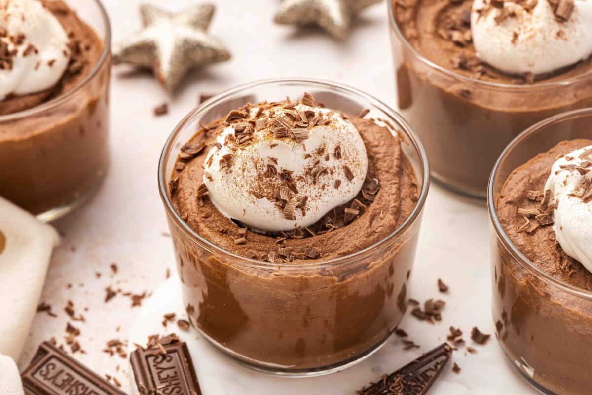 A glass dish of chocolate mousse topped with whipped cream and chocolate shavings, with more chocolate mousse desserts and star decorations in the background.