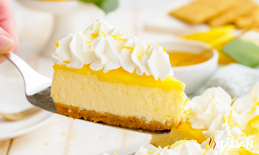 A slice of lemon cheesecake with whipped cream on top is being lifted from a whole cheesecake on a light-colored table.