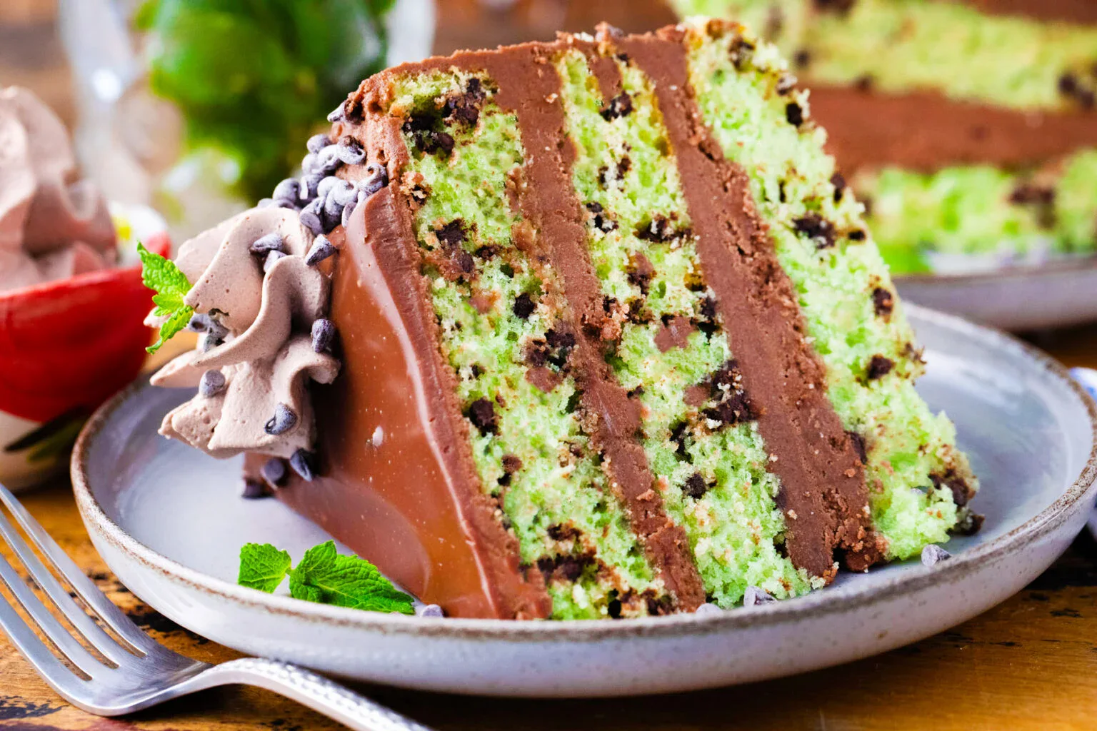 A slice of layered mint chocolate chip cake with chocolate frosting and chocolate chips, served on a plate with a fork and a garnish of mint leaves.