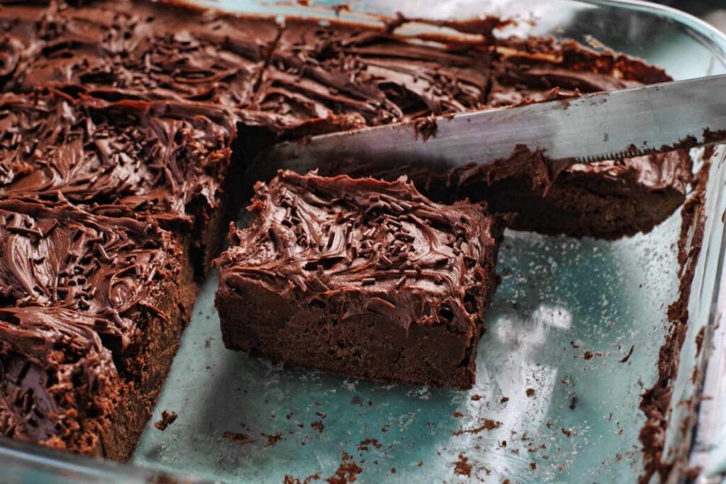 A knife lifting a frosted chocolate brownie from a glass baking dish with several other brownies inside.