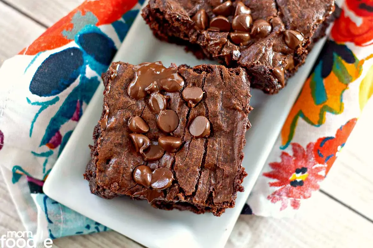 Two chocolate brownies topped with chocolate chips are placed on a white rectangular plate, which sits on a colorful floral-patterned napkin.