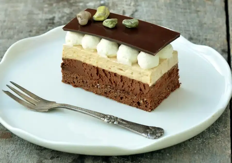 A slice of layered chocolate and cream dessert topped with whipped cream, pistachios, and a chocolate piece, served on a white plate with a vintage fork.