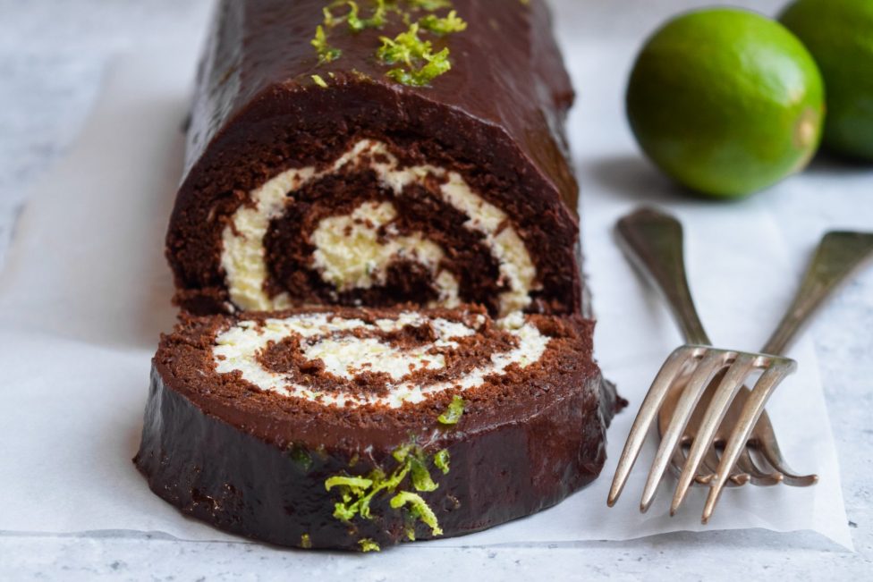 A chocolate Swiss roll cake with cream filling, topped with chocolate glaze and lime zest, sits on parchment paper near two forks and two whole limes.