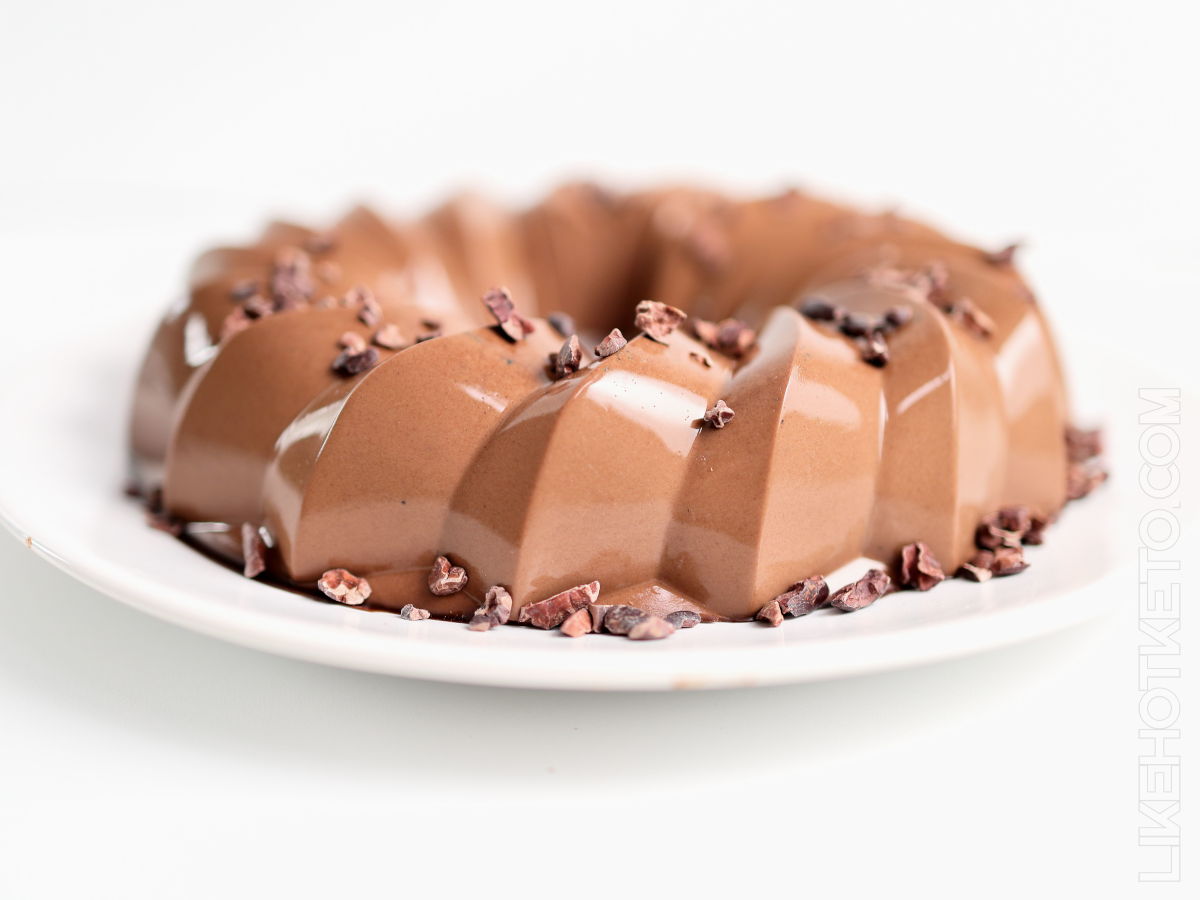 A chocolate mousse bundt cake topped with cacao nibs is displayed on a white plate against a white background.