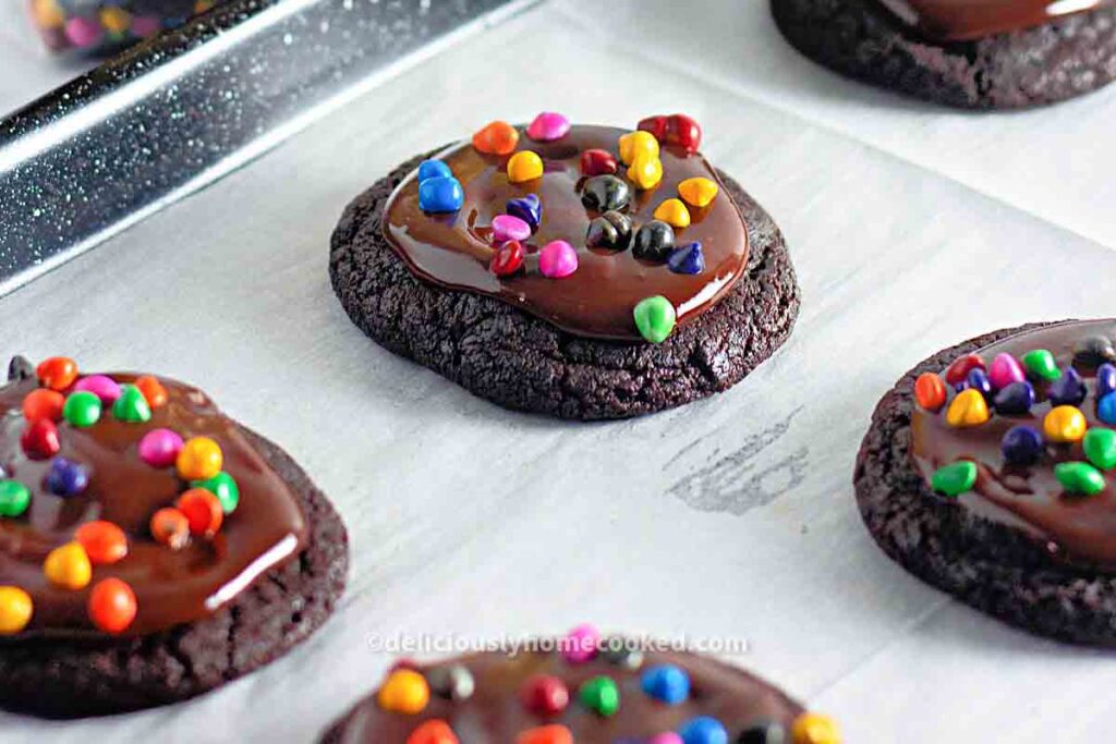 Chocolate cookies topped with a layer of chocolate frosting and colorful candy-coated pieces, arranged on parchment paper.