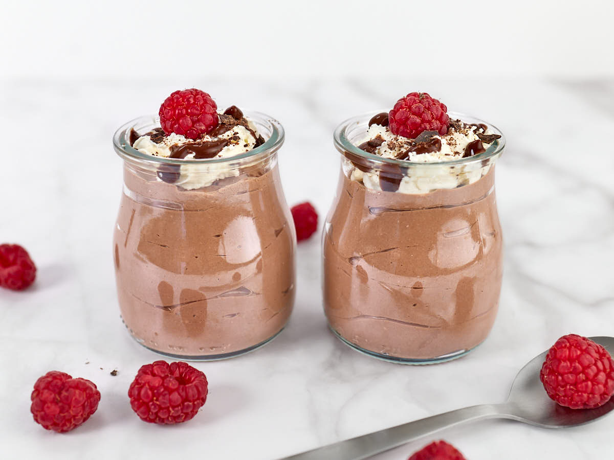 Two small glass jars filled with chocolate mousse, topped with whipped cream, chocolate drizzle, and a raspberry. Several loose raspberries and a spoon are nearby on a marble surface.