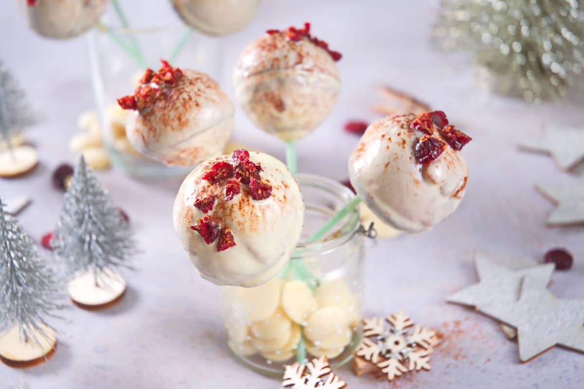 White chocolate cake pops on sticks, decorated with red dried fruit and dusted cocoa, displayed in glass jars with white chocolate pieces. Silver trees and snowflakes are in the background.