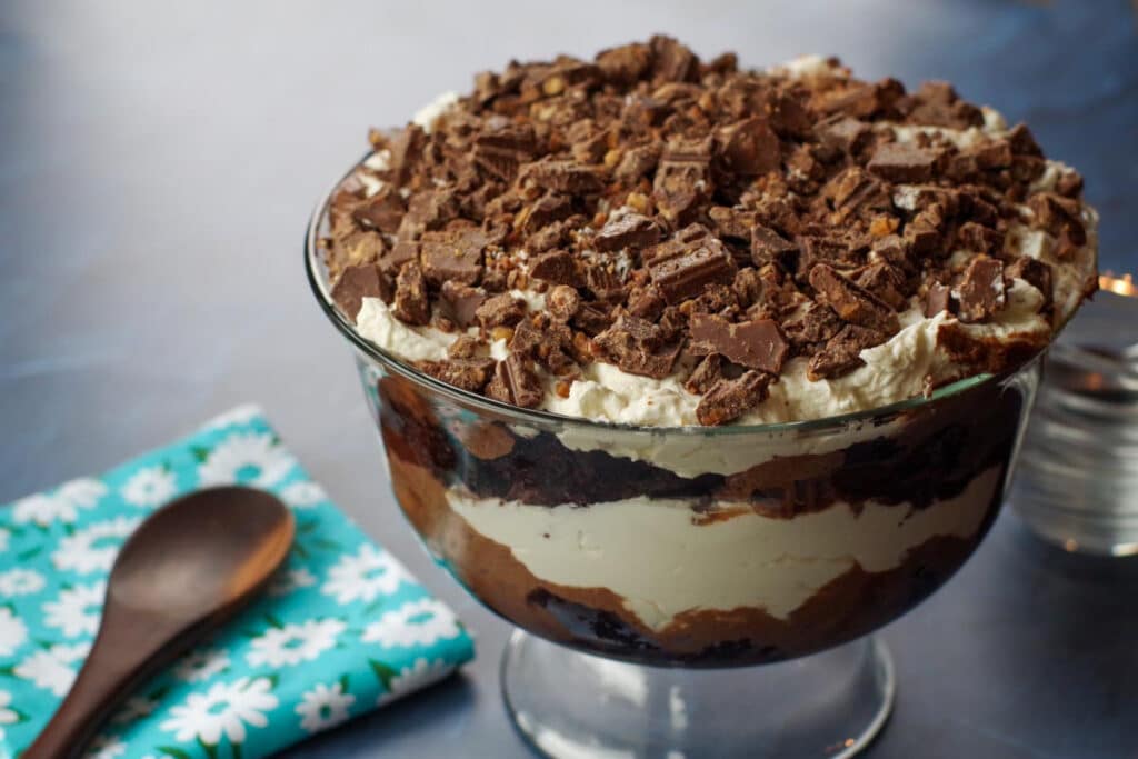 A glass trifle bowl filled with layered dessert of whipped cream, chocolate, and crumbled cookies, placed next to a blue floral napkin and a wooden spoon.