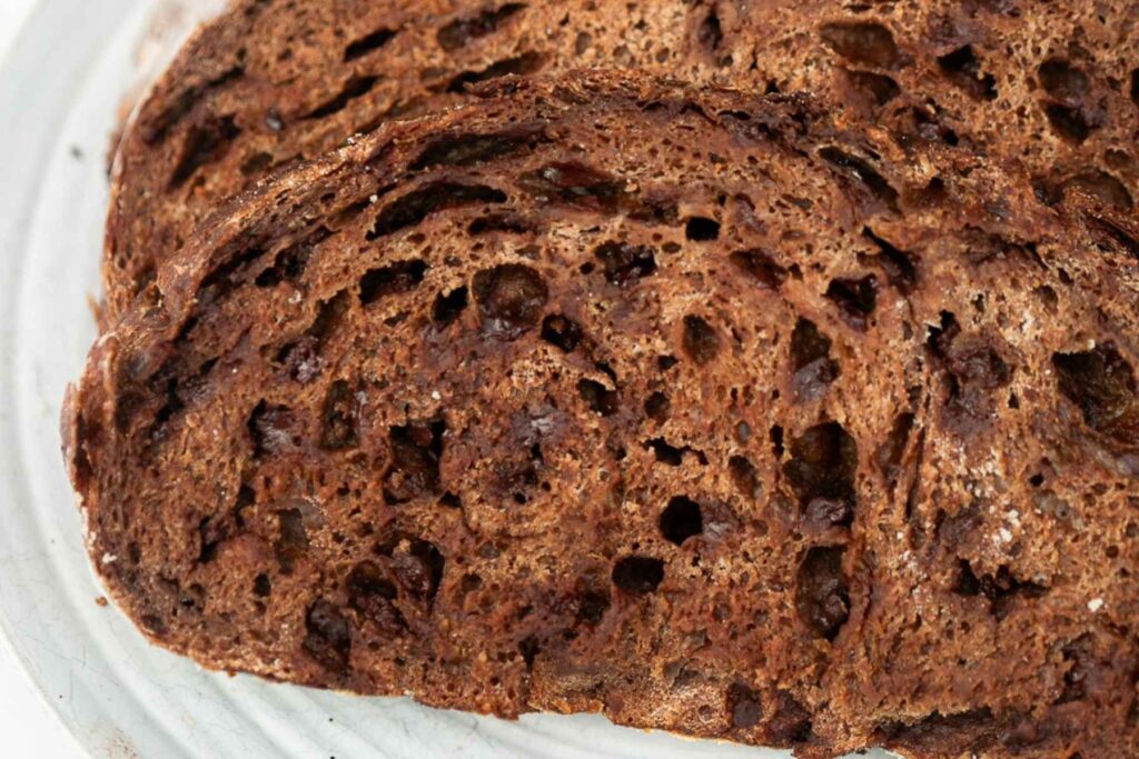 Two slices of dark, rustic bread with a porous, airy texture are shown on a white plate.