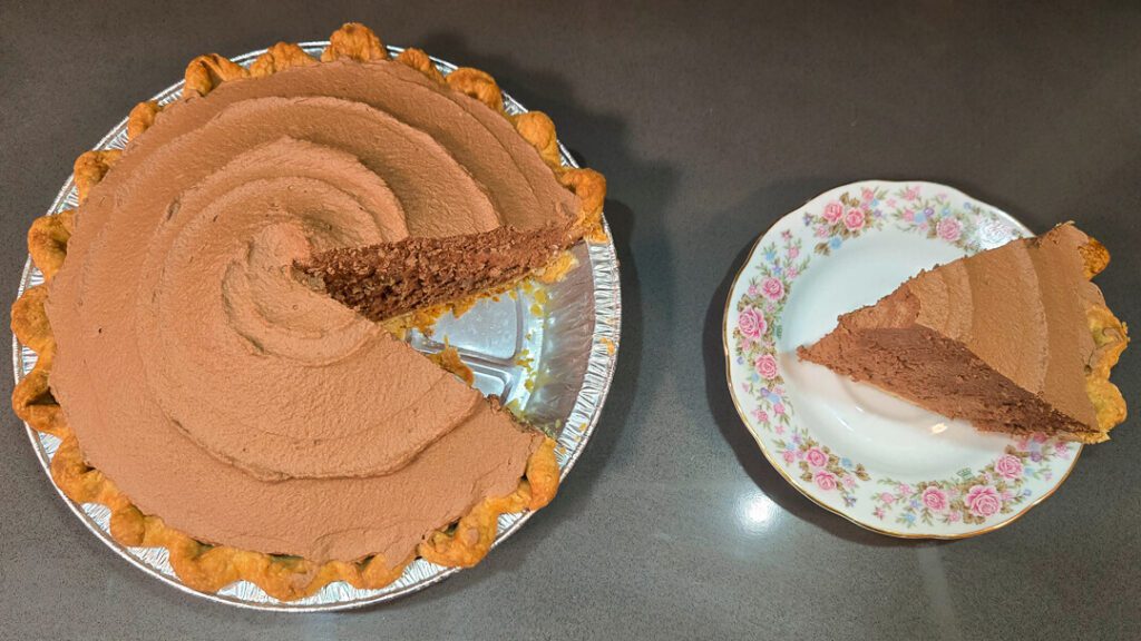 A chocolate cream pie with one slice removed; the slice is placed on a floral-patterned plate next to the pie.