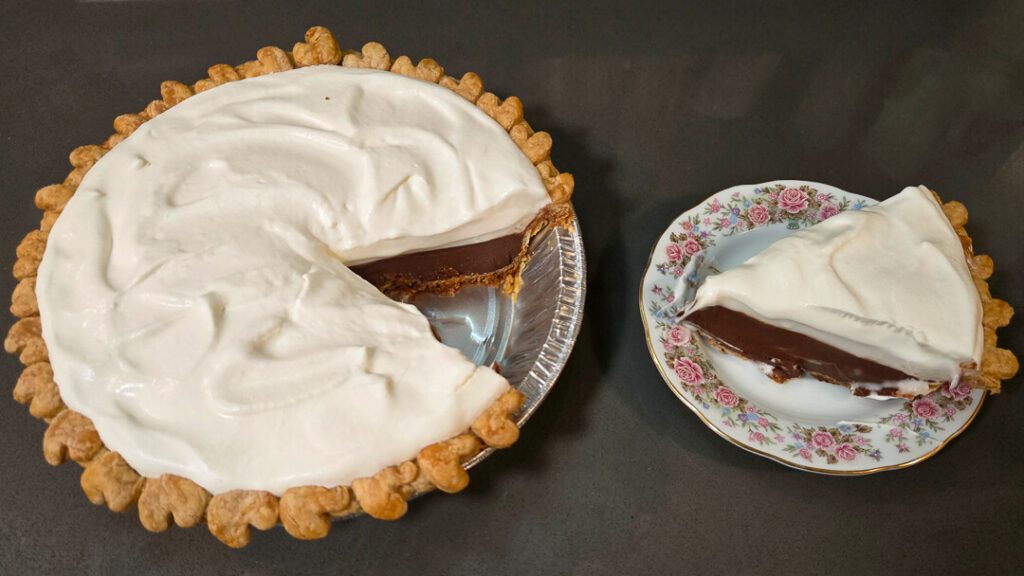 A chocolate cream pie with a slice removed sits next to a dessert plate holding the single slice, both topped with a layer of whipped cream.