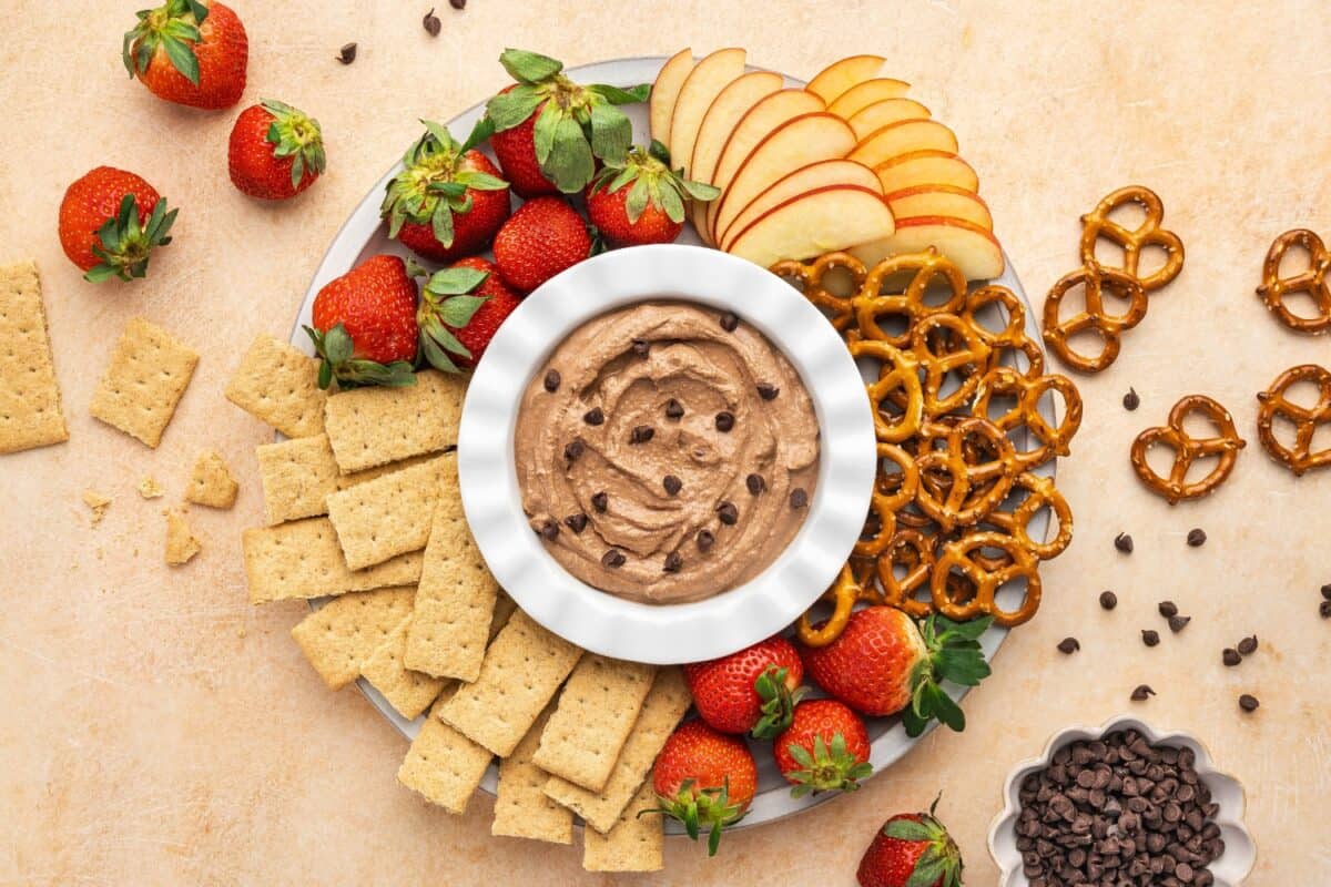 A plate with strawberries, apple slices, pretzels, and graham crackers arranged around a bowl of chocolate chip dip. Scattered pretzels and chocolate chips on the table.