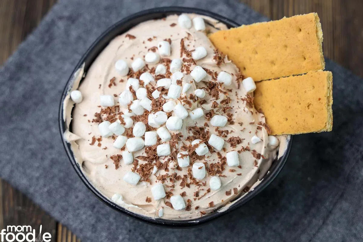 A bowl of whipped chocolate dessert topped with mini marshmallows, chocolate shavings, and two graham cracker pieces, placed on a dark cloth.