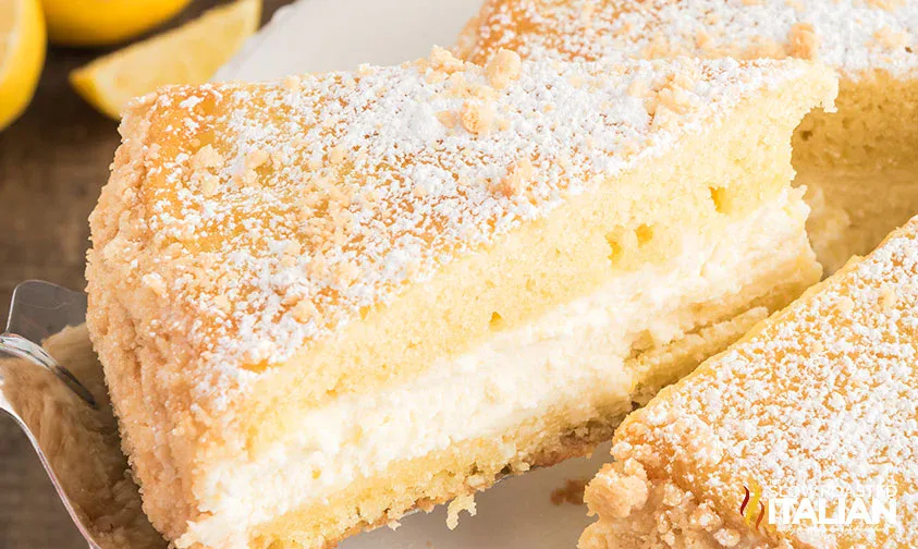 A slice of layered sponge cake with a creamy filling, topped with powdered sugar, is served on a plate. Lemons are visible in the background.