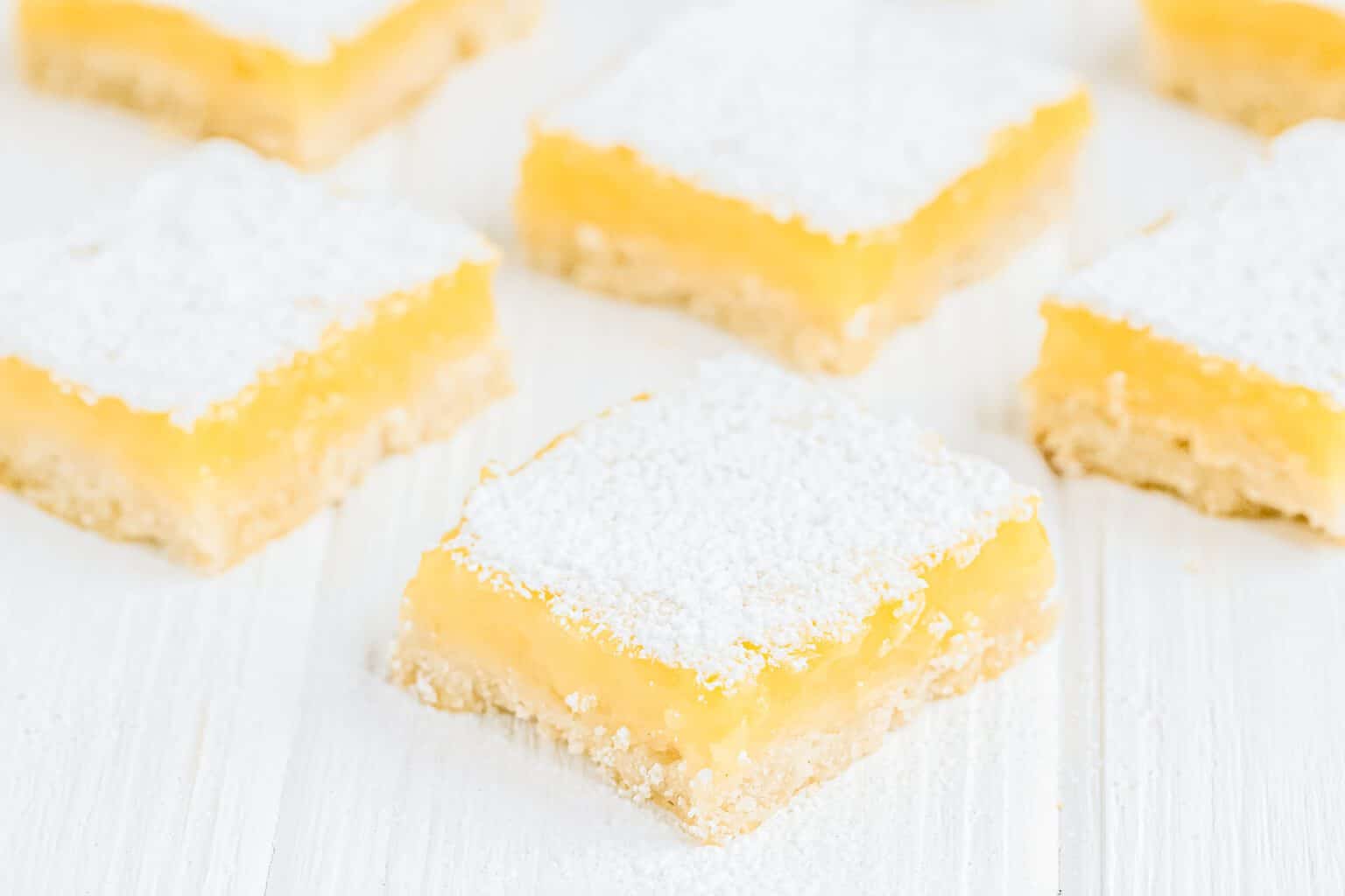 Several lemon bars topped with powdered sugar are arranged on a white surface.