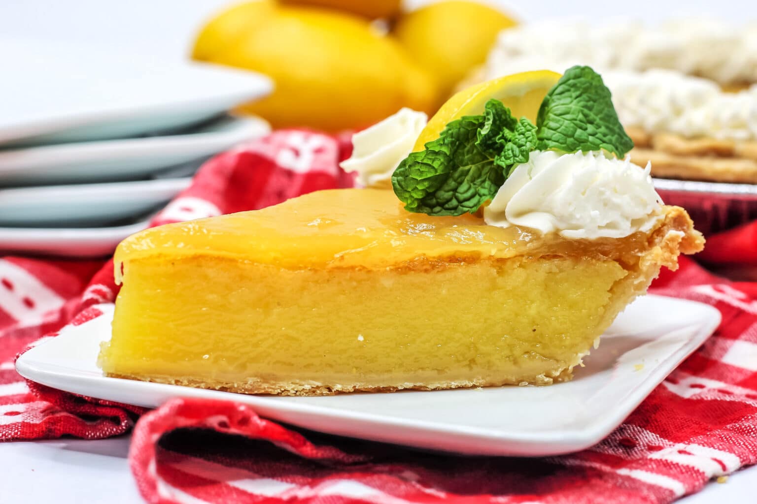 A slice of lemon pie garnished with whipped cream, a lemon slice, and mint leaves on a white plate, set on a red checkered cloth.