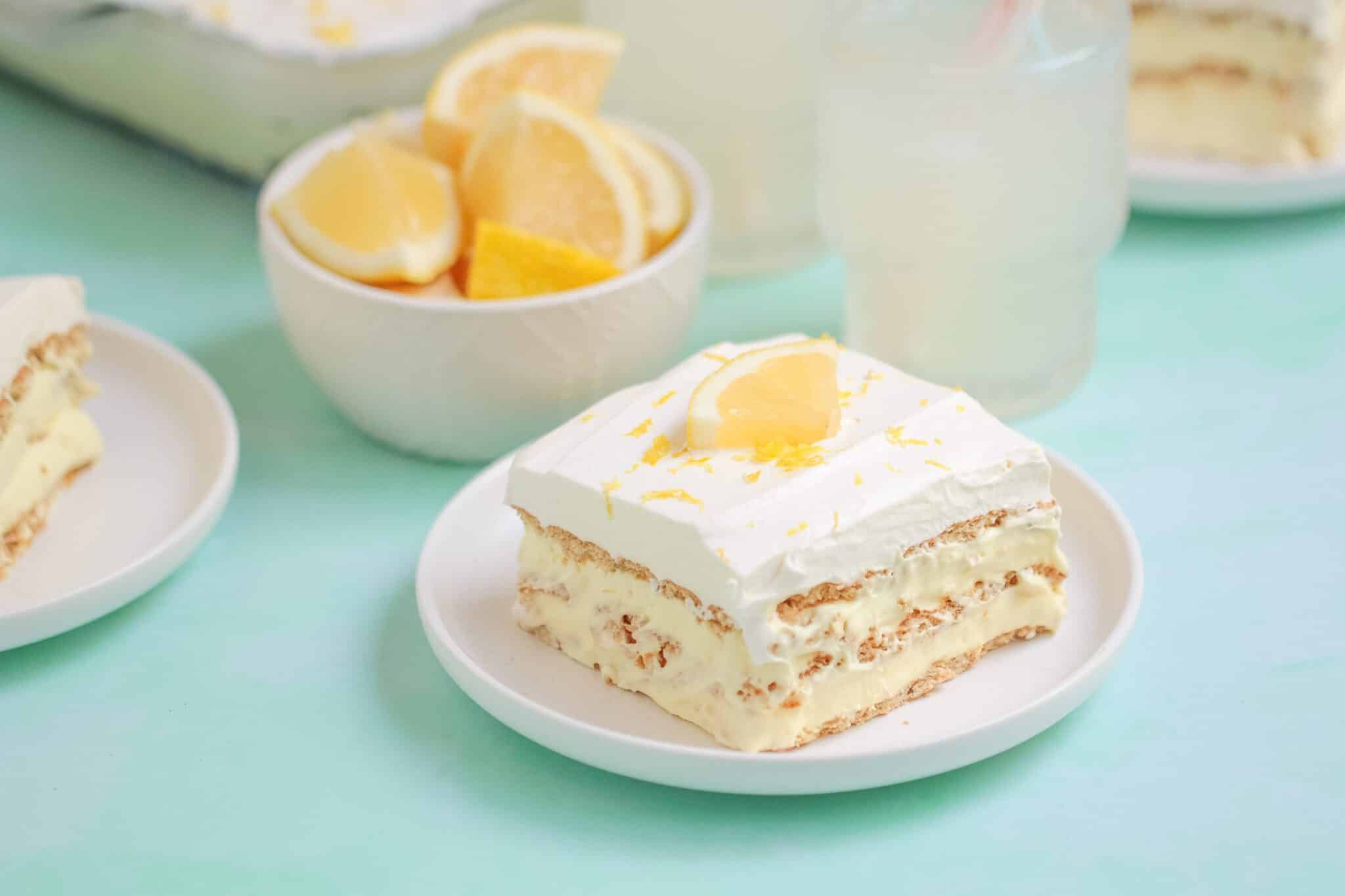 A slice of layered lemon dessert with whipped topping and a lemon wedge is served on a white plate; a bowl of lemon slices is in the background.