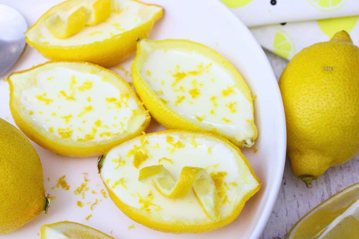 Lemon halves filled with creamy lemon dessert, topped with lemon zest, arranged on a white plate with whole lemons nearby.