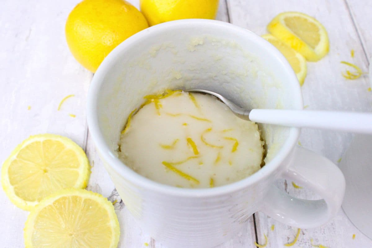 A white mug with lemon cake topped with icing and lemon zest, surrounded by fresh lemon slices and whole lemons on a white surface.