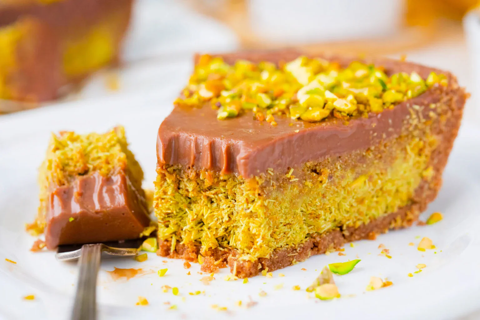 A slice of pistachio cake with a chocolate topping sits on a white plate, with a forkful of cake resting beside it.