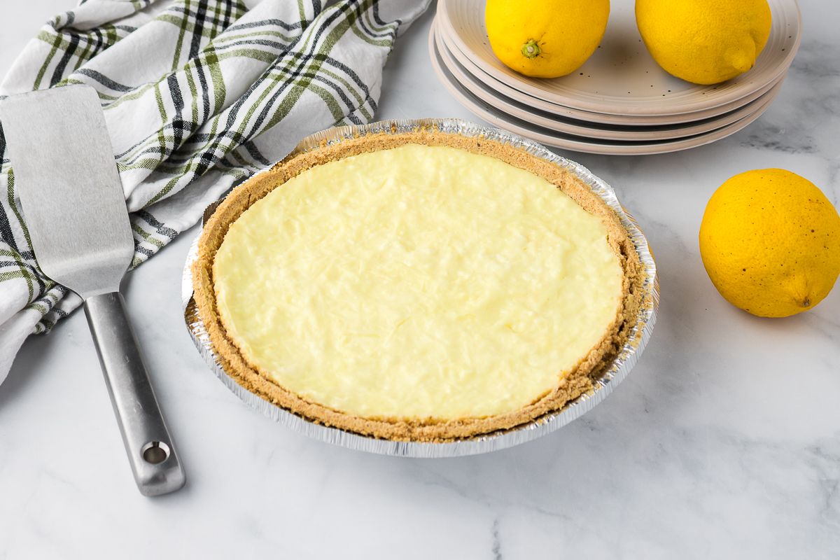 A lemon pie with a graham cracker crust sits on a marble countertop beside a metal spatula, a striped towel, three lemons, and a stack of plates.