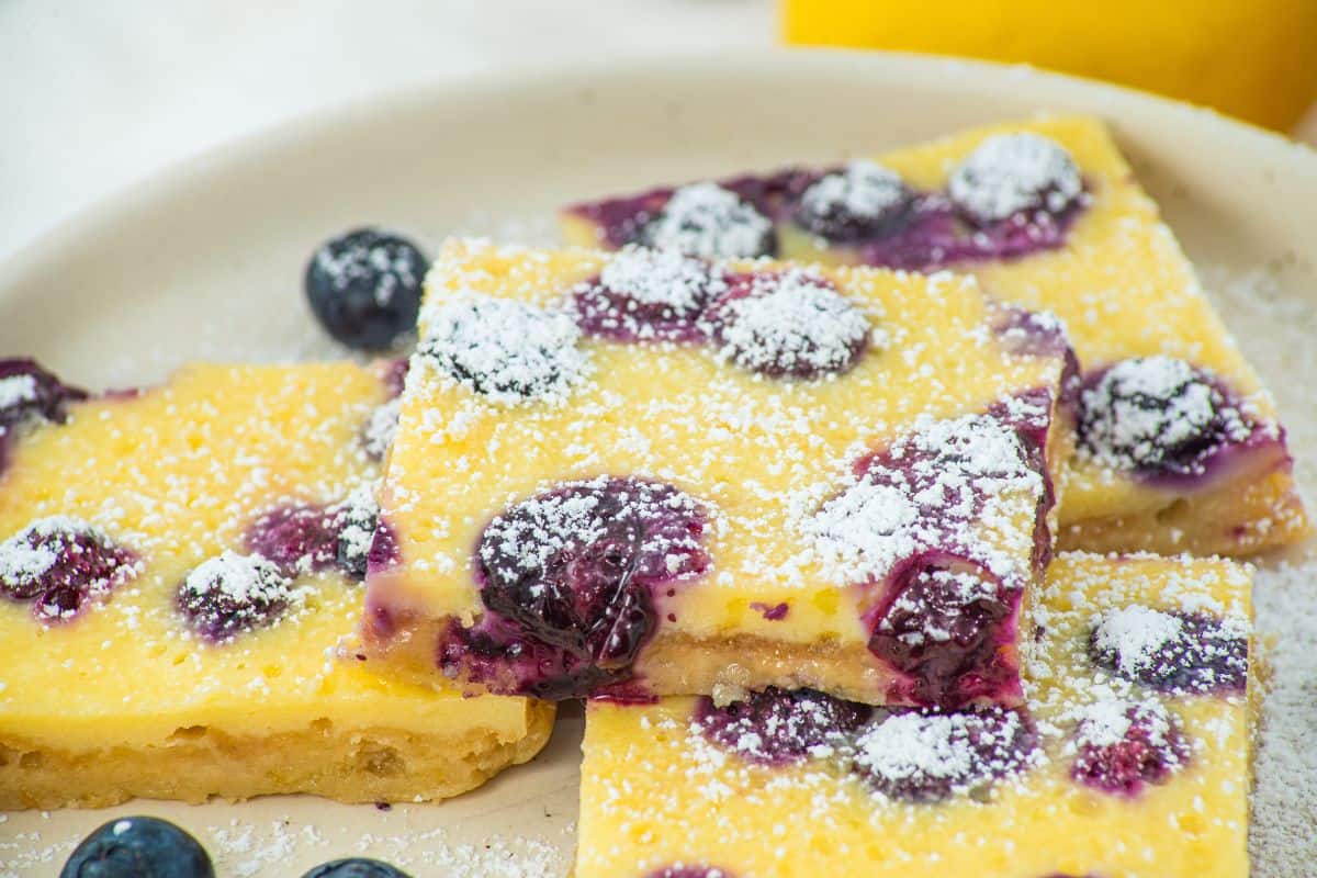Three lemon blueberry bars topped with powdered sugar are stacked on a plate, with a few fresh blueberries scattered around.