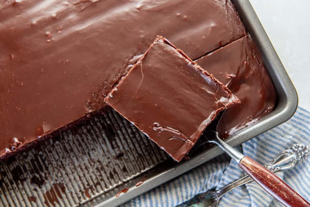 A metal spatula lifts a square piece of chocolate sheet cake with glossy chocolate frosting from a baking pan.