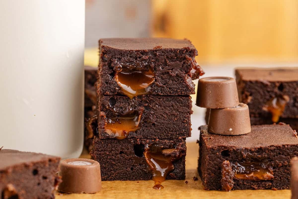 Stacked chocolate brownies with oozing caramel filling are placed next to a glass of milk and several round chocolate candies on a wooden surface.