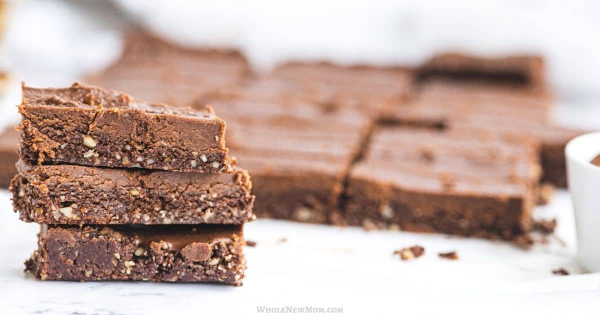 Stack of three chocolate brownies with visible nut pieces in front of a tray of neatly cut brownies on a white surface.