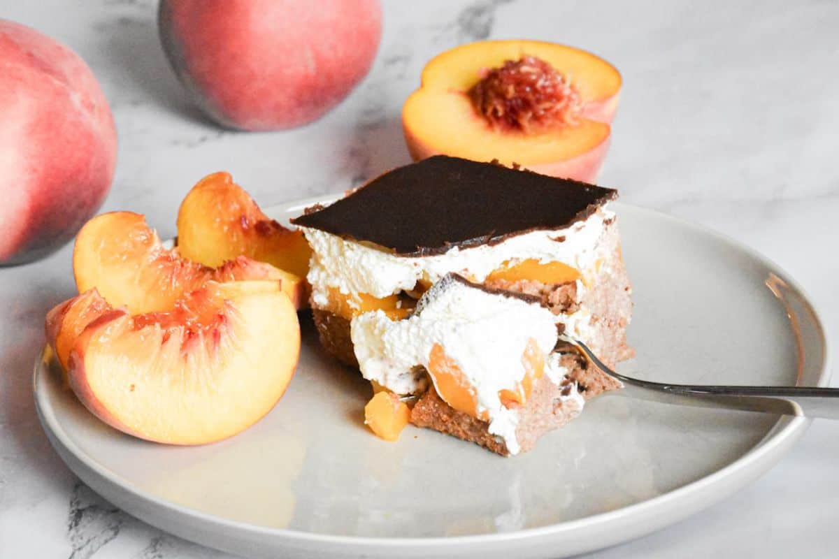 A slice of dessert with whipped cream, peaches, and a chocolate layer sits on a plate beside fresh peach slices and a halved peach.